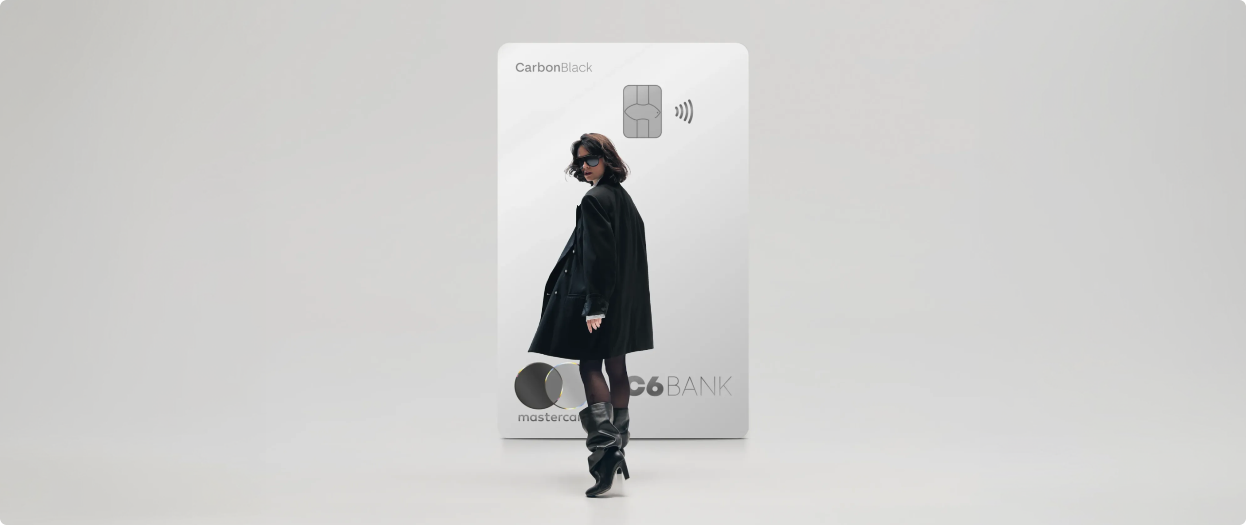 Mulher de preto caminha à frente de um Cartão C6 Carbon Mastercard Black prata em tamanho ampliado, sobre fundo claro.